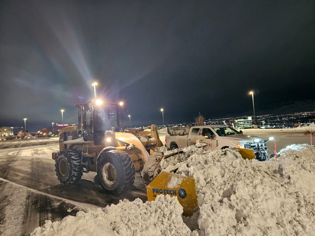 Heavy machinery clearing snow at night in a parking lot.