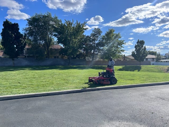 Landscaping crew mowing a green Saratoga Springs lawn with a riding mower under blue skies.